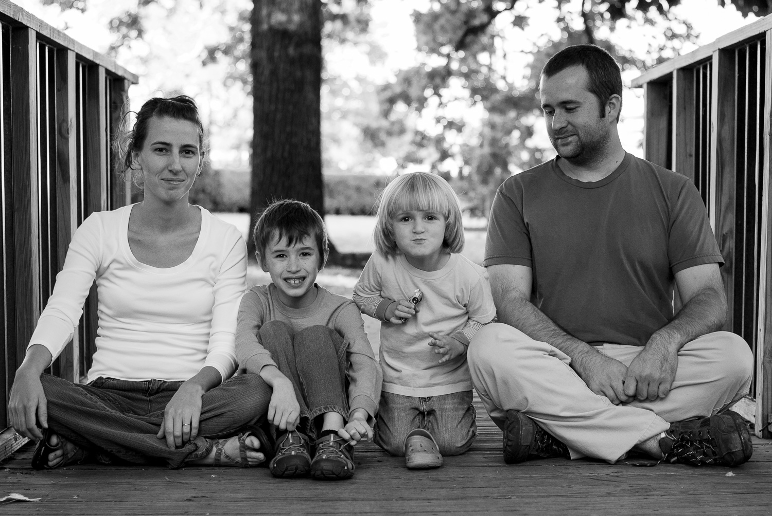 Black and white family photos. Two parents and their young children sitting cross-legged on a walking bridge.